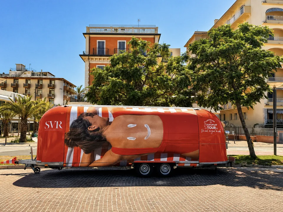 Caravane Airstream habillée SVR d'une baigneuse en maillot rayé, stationnée sur une place bordée de palmiers